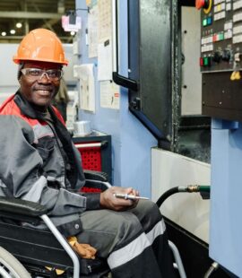 An employee in a hardhat, protective eyewear, and wheelchair who is smiling while on the clock because his employer is making him feel empowered this National Disability Employment Awareness Month.